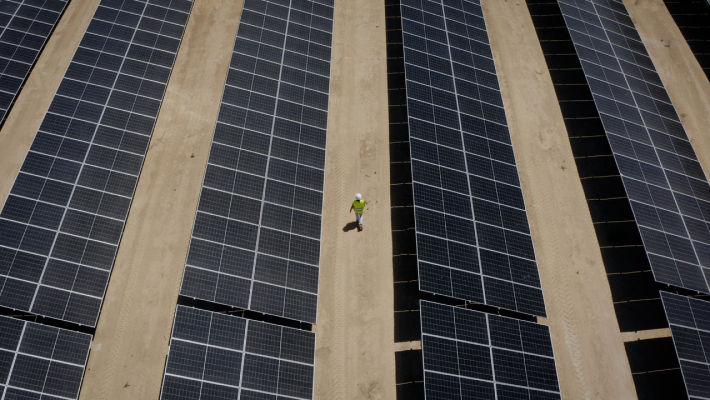 solar farm with rows of panels on sandy ground and a person in a green shirt and hard hat walking between them