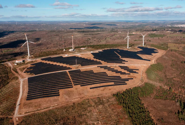 hybrid renewable energy site with rows of solar panels and three wind turbines in the background, set in a sparse landscape with patches of greenery