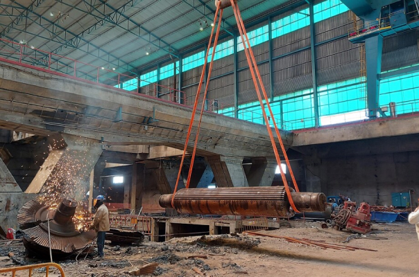 interior view of a thermal power station undergoing reconversion. the image features industrial equipment, including a large cylindrical structure being lifted by orange straps attached to a crane. workers are visible, one using a tool emitting sparks, indicative of cutting or welding metal. the space has high ceilings, metal beams, and large windows, with scattered debris and machinery