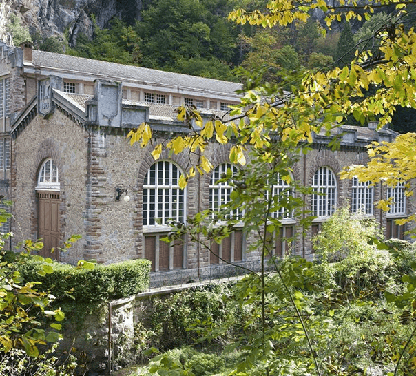 stone building with arched windows and wooden doors, surrounded by lush greenery, including trees, bushes, and plants. a rocky cliff in the background adds to the natural setting