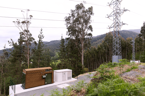 rural electrical substation with a wooden structure, a green door, and a white utility box on a concrete platform. power lines and electrical towers extend across the landscape, surrounded by trees and hills in the distance