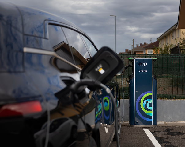 an electric vehicle (ev) is being charged at an edp charge station, which features the edp logo and 'charge' branding. the station is in a residential or suburban area, with houses and a wooden fence visible in the background