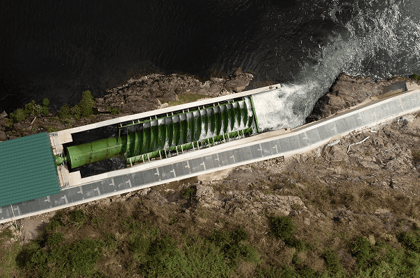 aerial view of a hydroelectric power plant featuring a green archimedes screw turbine within a concrete structure. water flows through the turbine, exiting into a river, with rocky terrain and vegetation surrounding the site