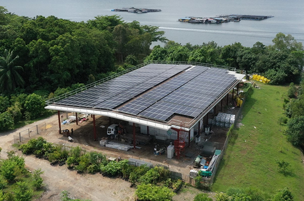 a large industrial building with solar panels covering its roof, located near a green field and a body of water. surrounding the building are various industrial materials and equipmen