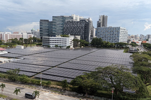  large field of solar panels arranged in a grid pattern, set within an urban area. the background features a variety of buildings of different styles and heights