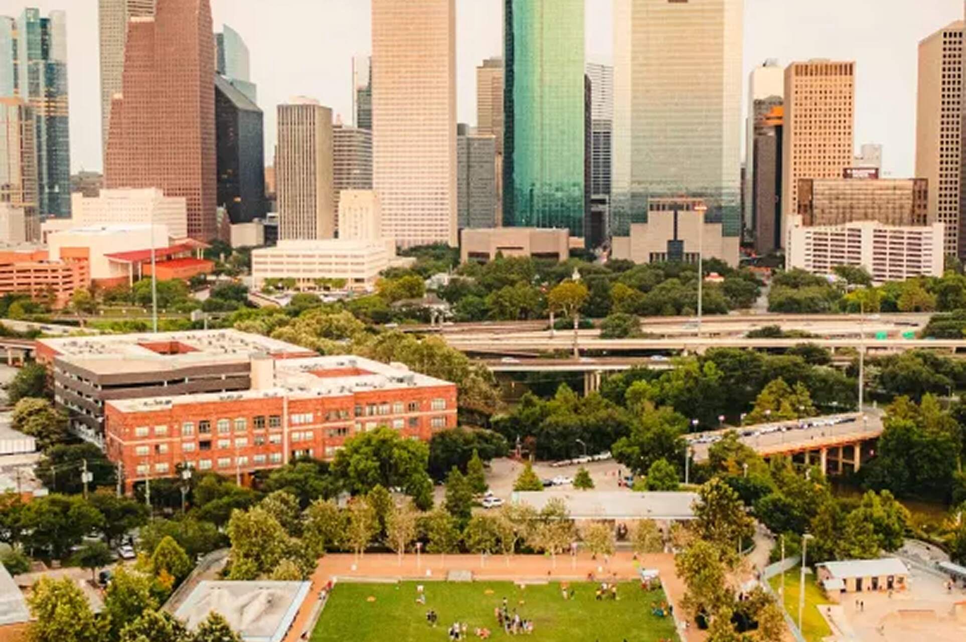 red brick building surrounded by trees and with soccer field in front. in the background, a highway and a skyline filled with skyscrapers