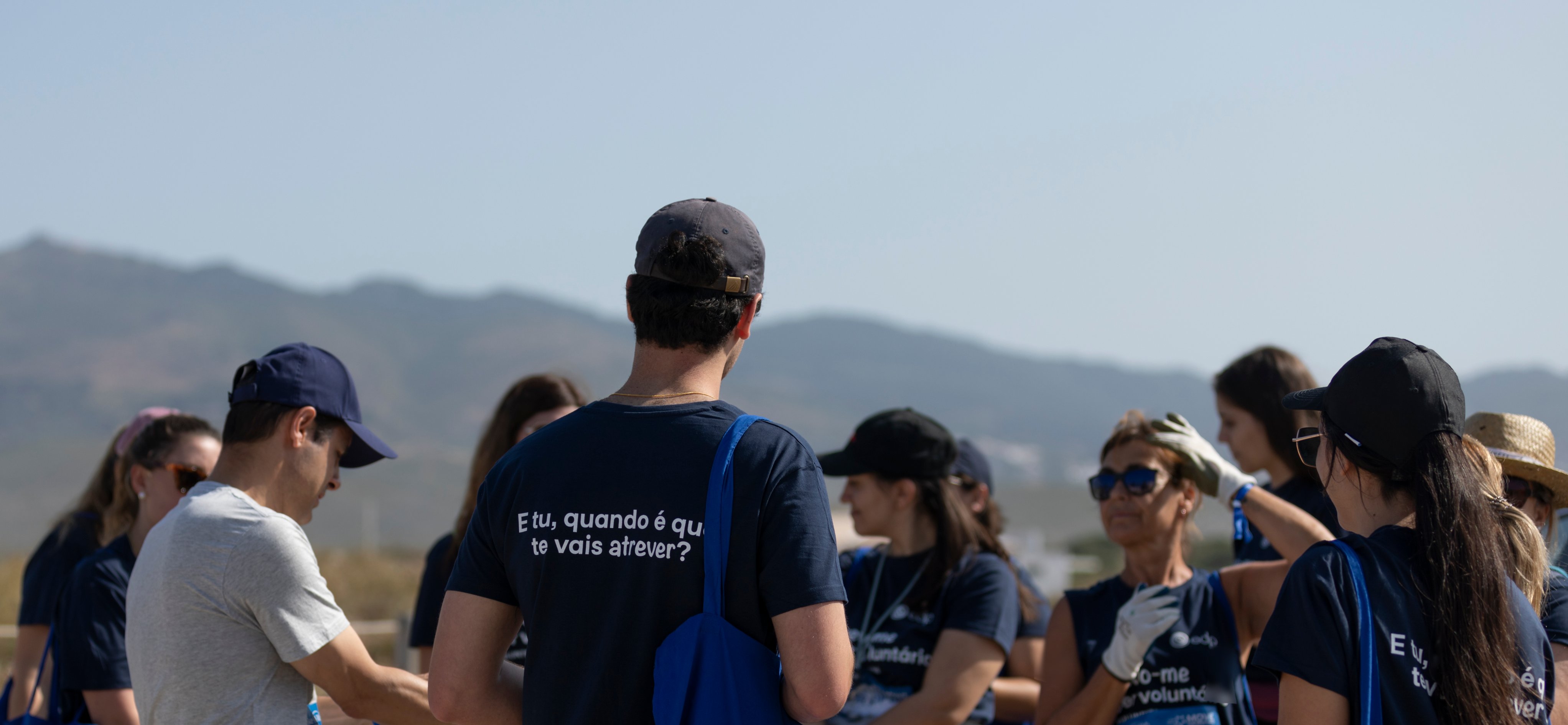 group of employees gathered on the beach with one employee's back facing the camera wearing an official volunteering t shirt