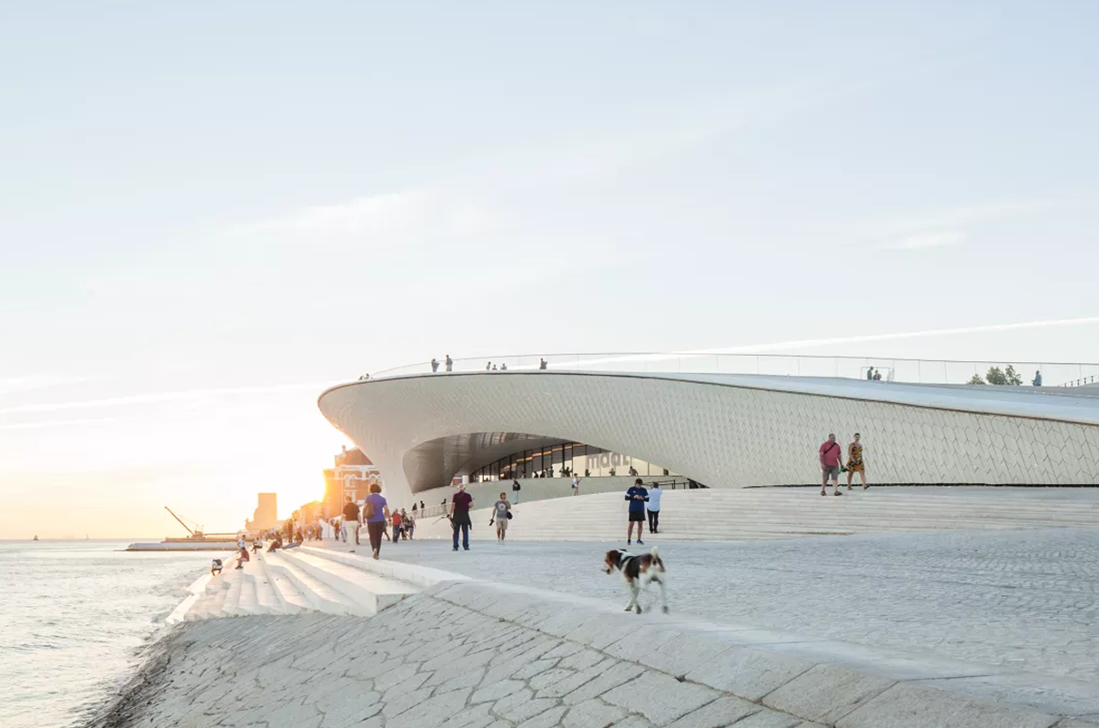 modern architectural view of the maat museum in lisbon, portugal, featuring its curved, textured facade along the tagus river waterfront. people are seen walking on the promenade and rooftop, enjoying the scenic river backdrop under a serene sky