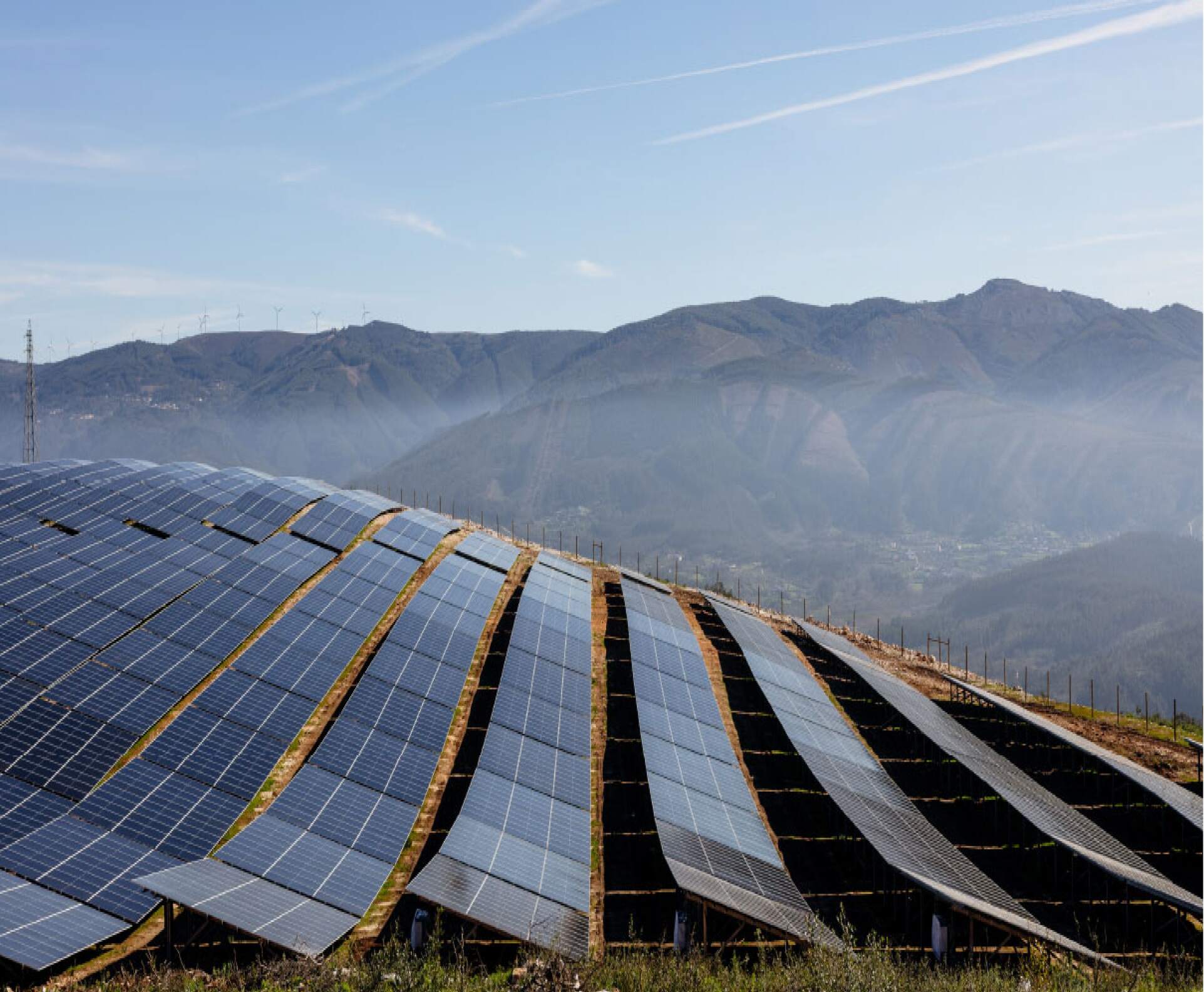 solar panels arranged in rows on a hillside, with mountains in the background under a partly cloudy sky