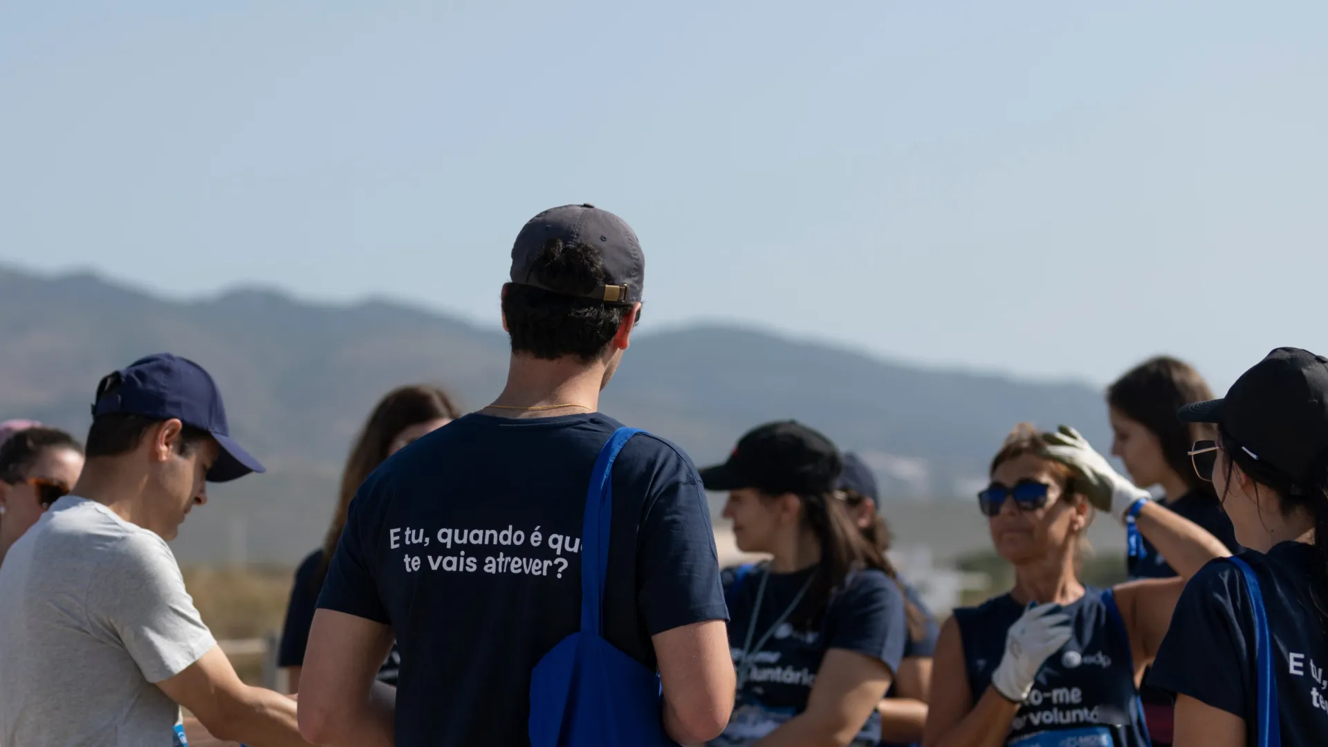 group of employees gathered on the beach with one employee's back facing the camera wearing an official volunteering t shirt 